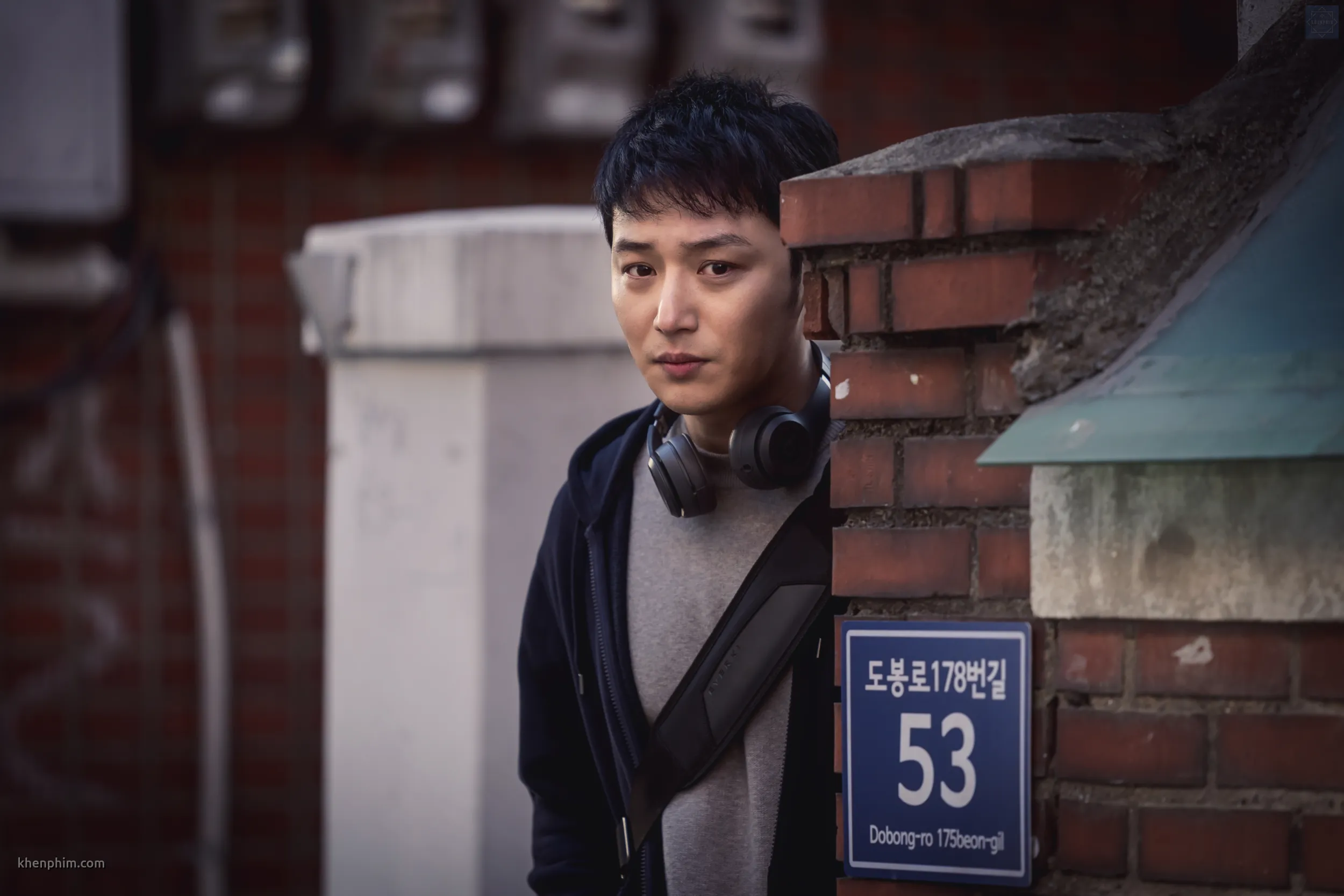 Koo Jung Tae (Byun Yo Han) lấp ló sau bức tường với ánh mắt ám ảnh, dõi theo ai đó. Anh ăn vận đơn giản, đeo tai nghe và mang theo một chiếc balo. Biểu cảm của Jung Tae toát lên vẻ bí ẩn, khó đoán, phản ánh nội tâm phức tạp của một kẻ bám đuôi.
